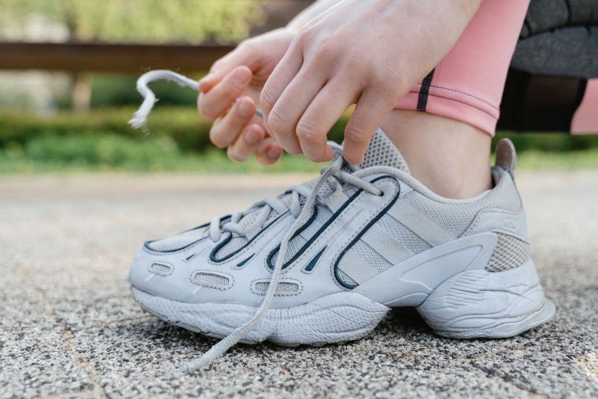 Hands tying the laces of a gray sneaker on a paved path.